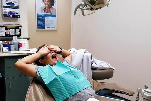 A boy covering his eyes while lying on a dentist chair in a dentist office.