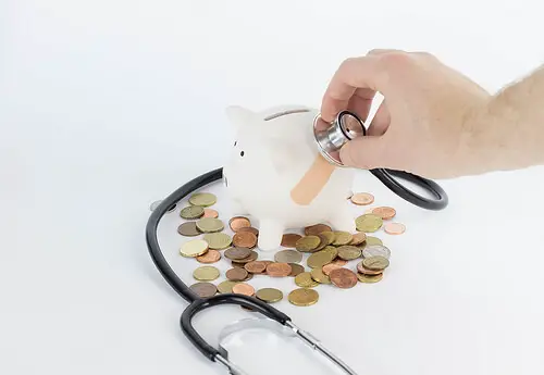 A hand using a stethoscope on a white piggy bank with coins scattered.